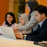 Sekelompok profesional muda berdiskusi di kantor menggunakan laptop sebagai ilustrasi hard skill dan soft skill di dunia kerja.